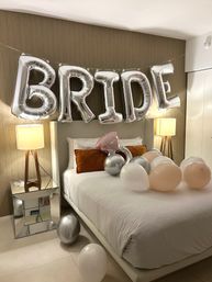 Silver foil balloons spelling "BRIDE" above a modern bridal suite bed with white linens, peach and silver balloons, mirrored nightstand and bedside lamps — wedding prep decor.