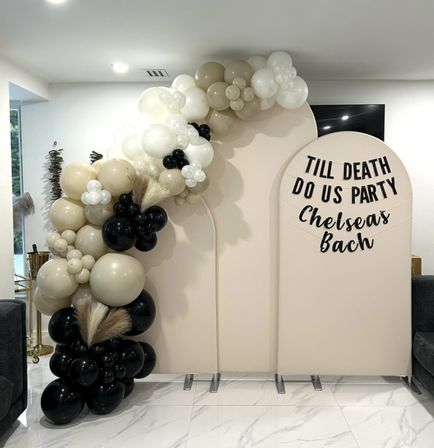 Neutral beige, cream, white and black balloon garland with pampas grass framing two arched blush backdrops in a modern indoor living room, one featuring a playful "Till death do us party" bachelorette banner.