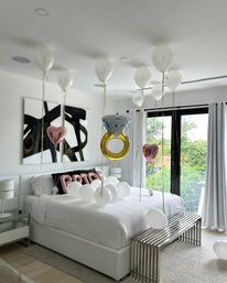 Sunlit modern bedroom with white bedding and sliding glass doors, decorated for a celebration with floating white balloons, rose-gold heart balloons, a large gold engagement-ring balloon, and rose-gold letter balloons on the bed.