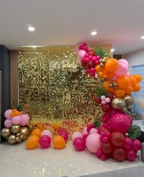 Indoor party photo backdrop with a shimmering gold sequin wall and neon "Let's Party" sign, styled with a vibrant pink, orange, burgundy and gold balloon garland and tropical leaves.