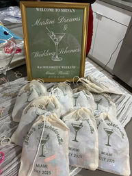 Gold-framed green sign for a martini-themed bachelorette weekend in Miami (July 2025) displayed next to matching drawstring favor bags printed with martini glass icons on a table.