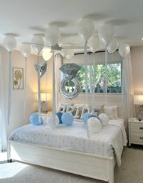 Bright coastal-style bedroom decorated for a celebration with white and pale-blue helium balloons, a large silver engagement-ring foil balloon and silver letter balloons on the headboard, soft natural light from a window.