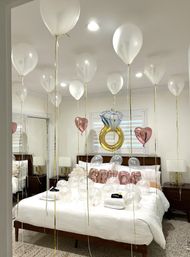 Home bridal surprise: white helium balloons with gold ribbons float above a white-made bed scattered with clear balloons, rose-gold heart accents, 'BRIDE' letter balloons and a gold engagement-ring foil balloon.