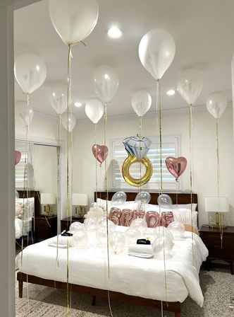 Home bridal surprise: white helium balloons with gold ribbons float above a white-made bed scattered with clear balloons, rose-gold heart accents, 'BRIDE' letter balloons and a gold engagement-ring foil balloon.