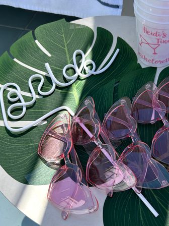 Stack of pink heart-shaped sunglasses on a green monstera leaf placemat with a white decorative wire sign and a party cup — poolside bachelorette favors.