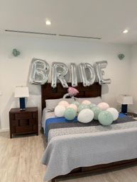 Bridal bedroom decorated with large silver 'BRIDE' letter balloons above a wooden headboard and pastel green, pink and white balloons scattered on a gray quilted bed in a modern room with wood flooring and bedside lamps