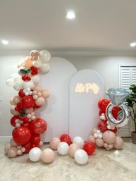 Indoor party backdrop with white arched panels, blush-red-white balloon arch and scattered balloons, small cherry-shaped balloons, glowing "Let's Party" neon sign and a large diamond-ring foil balloon on tiled floor.