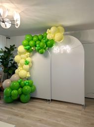 Bright green and lemon-yellow balloon garland draped over a white arched photo backdrop with a glowing "Let's Party" neon sign in a cozy living-room setting