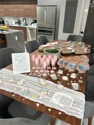 Mermaid-themed bachelorette party spread on a wooden dining table in a modern home kitchen, with pink champagne flutes, decorated sugar cookies, woven gift bags and seashell accents.