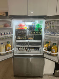 Open stainless steel French-door refrigerator in a modern kitchen, neatly stocked with bottled water, orange juice, canned beverages in a lower drawer, wine and soda bottles, and containers of pre-cut fruit and prepared salads