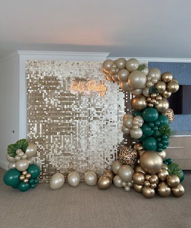 Indoor gold sequin event backdrop with neon Lets Party sign and large green and metallic gold balloon garland accented by leopard-print balloons and tropical leaves