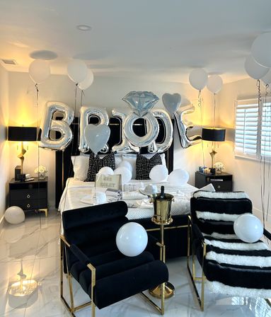 Modern black-and-white bedroom set up for a bridal surprise with giant silver letter and diamond-ring balloons, white heart and round balloons, made bed with patterned pillows, gold-accent chairs and champagne bucket, bedside lamps, and glossy tile floor.