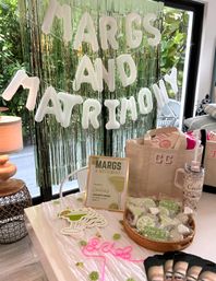Margarita-themed bachelorette party display in Miami, FL — white balloon letters reading MARGS AND MATRIMONY over green foil fringe backdrop, lime cookies, tote bags, tumblers and neon celebrate sign.