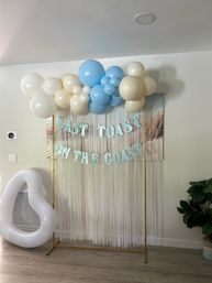 Playful indoor coastal-themed celebration backdrop with cream and light-blue balloon garland over a white fringe curtain and glitter letters reading LAST TOAST ON THE COAST, flanked by an inflatable prop and potted plant.