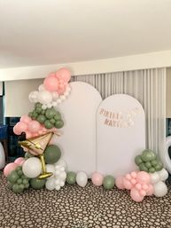 Indoor photo-booth party backdrop with two white arched panels, pastel pink, white and olive balloon garland, gold martini foil balloon and banner reading "Bikinis and Martinis"