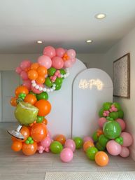Cheerful indoor party photo backdrop with a white arch, neon 'Let's Party' sign, and a vibrant balloon garland in pink, orange and green with a foil margarita balloon on a light wood floor.