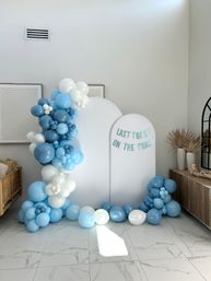 Playful coastal-themed photo backdrop with white arched panels reading “Last Toast on the Coast,” surrounded by blue and white balloon garlands in a modern room with marble tile floor and dried pampas grass.