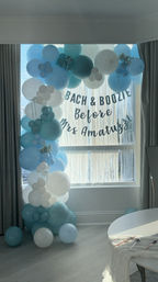 Blue and white balloon arch framing a bright window with metallic tassel fringe and a playful bride-to-be bachelorette party banner.