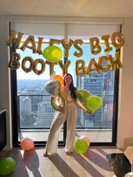 Smiling woman in a sunlit high-rise apartment holding a giant inflatable ring and lime balloons beneath gold foil letter balloons spelling a bachelorette party message, city skyline visible through the floor-to-ceiling window.