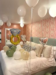 Bedroom celebration setup with white, gold, olive and silver balloons, large diamond ring foil balloon and heart-shaped balloons floating above a neatly made bed with green headboard and pink art-deco wallpaper