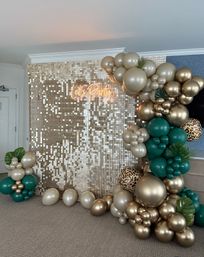 Indoor celebration backdrop with shimmering gold sequin wall and neon 'Let's Party' sign, framed by a metallic gold and emerald-green balloon garland with leopard-print accents and tropical leaves.