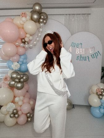 Smiling woman in white wearing a tiara and sash at an indoor bridal shower/bachelorette party with pastel pink, blue and gold balloon arch and a playful backdrop reading me? a bride? shut up!