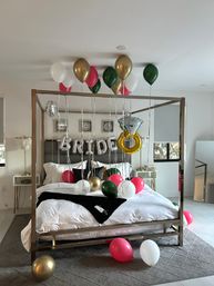 Modern canopy bed in a master bedroom decorated for a bridal surprise with gold, green, pink and white balloons, silver “BRIDE” letter balloons and a giant diamond ring balloon over white bedding.