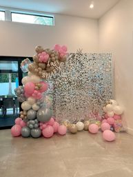 Indoor photo backdrop with pink, gray and gold balloon arch and clusters framing a sparkling silver sequin wall, pastel balloons scattered on a tiled floor