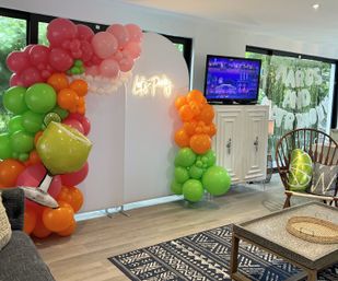 Indoor party backdrop with neon "Let's Party" sign framed by pink, orange and green balloon garlands, lime-shaped foil balloon, TV on white cabinet and cozy seating area with patterned rug.