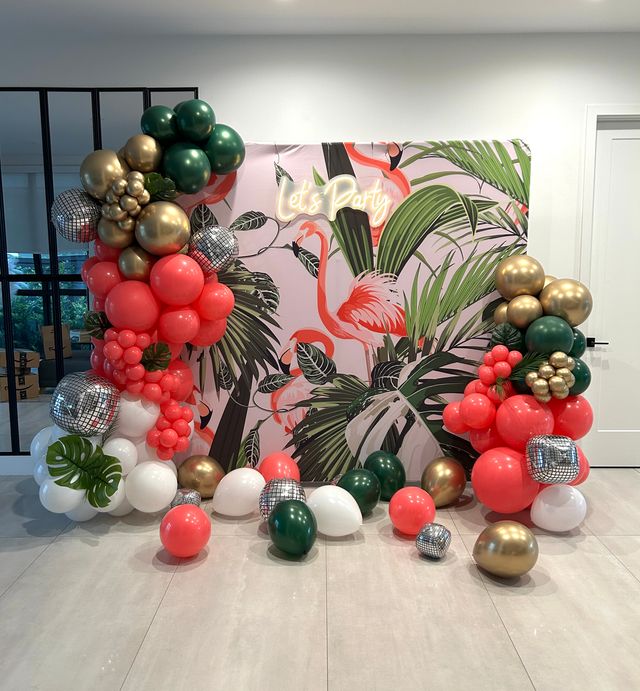 Indoor tropical flamingo party backdrop with "Let's Party" neon sign, framed by coral, dark green, white and metallic gold balloon garlands, mirrored disco-ball accents and scattered balloons on tiled floor.