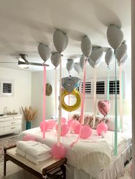 Bright white bedroom decorated for a romantic surprise with silver heart foil balloons tied to the ceiling by pastel ribbons, pink latex balloons scattered on the bed, and a large gold engagement-ring balloon centerpiece.