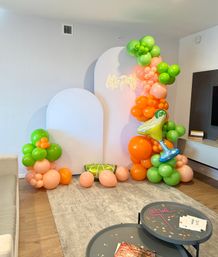 Indoor living room party backdrop with white arch panels and neon "Let's Party" sign, vibrant citrus balloon garland in green, orange and peach, inflatable cocktail and lime cushion, scattered balloons on rug.