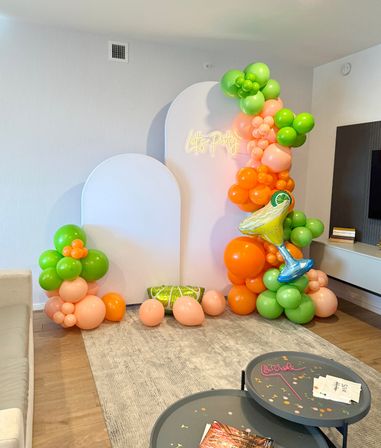 Indoor living room party backdrop with white arch panels and neon "Let's Party" sign, vibrant citrus balloon garland in green, orange and peach, inflatable cocktail and lime cushion, scattered balloons on rug.