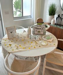 Sunlit modern dining nook set for a margarita bachelorette: tequila bottle piñata, paper cups, favor bags, white vases, tropical greenery and gold confetti on a draped round table.