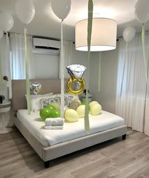 Modern neutral bedroom decorated for a playful bridal surprise with white and green balloons, a gold diamond-ring balloon, silver heart and "BRIDE" letter balloons, and folded towels on the bed.