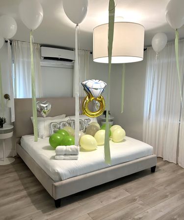 Modern neutral bedroom decorated for a playful bridal surprise with white and green balloons, a gold diamond-ring balloon, silver heart and "BRIDE" letter balloons, and folded towels on the bed.