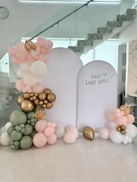 Indoor pastel party backdrop with blush pink, sage green and metallic gold balloon clusters framing white arched panels and a "Last Splash" banner beneath a glass staircase