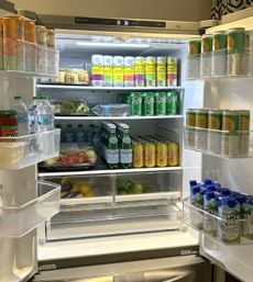 Open stainless-steel kitchen fridge packed with colorful canned seltzers, sparkling water, bottled water, soda, cut fruit trays and fresh produce.