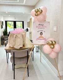 Modern dining room staged for a bachelorette party with pink-and-gold balloon clusters, a welcome sign on an easel, and canvas gift bags filled with pink tissue on a wooden table, light-filled interior with garden view.