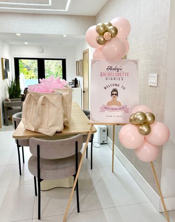 Modern dining room staged for a bachelorette party with pink-and-gold balloon clusters, a welcome sign on an easel, and canvas gift bags filled with pink tissue on a wooden table, light-filled interior with garden view.