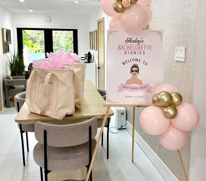 Modern dining room staged for a bachelorette party with pink-and-gold balloon clusters, a welcome sign on an easel, and canvas gift bags filled with pink tissue on a wooden table, light-filled interior with garden view.