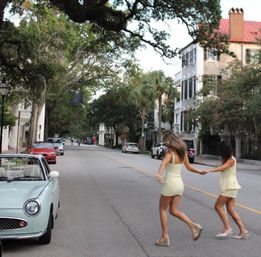 Two women in light summer dresses holding hands as they skip across a quiet oak- and palm-lined residential street past a mint vintage car and pastel townhouses