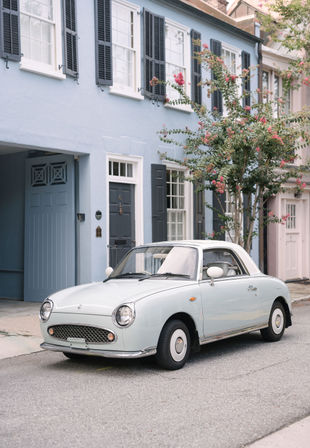 Pale blue vintage convertible parked on a quiet street in front of pastel row houses with black shutters and a flowering tree.