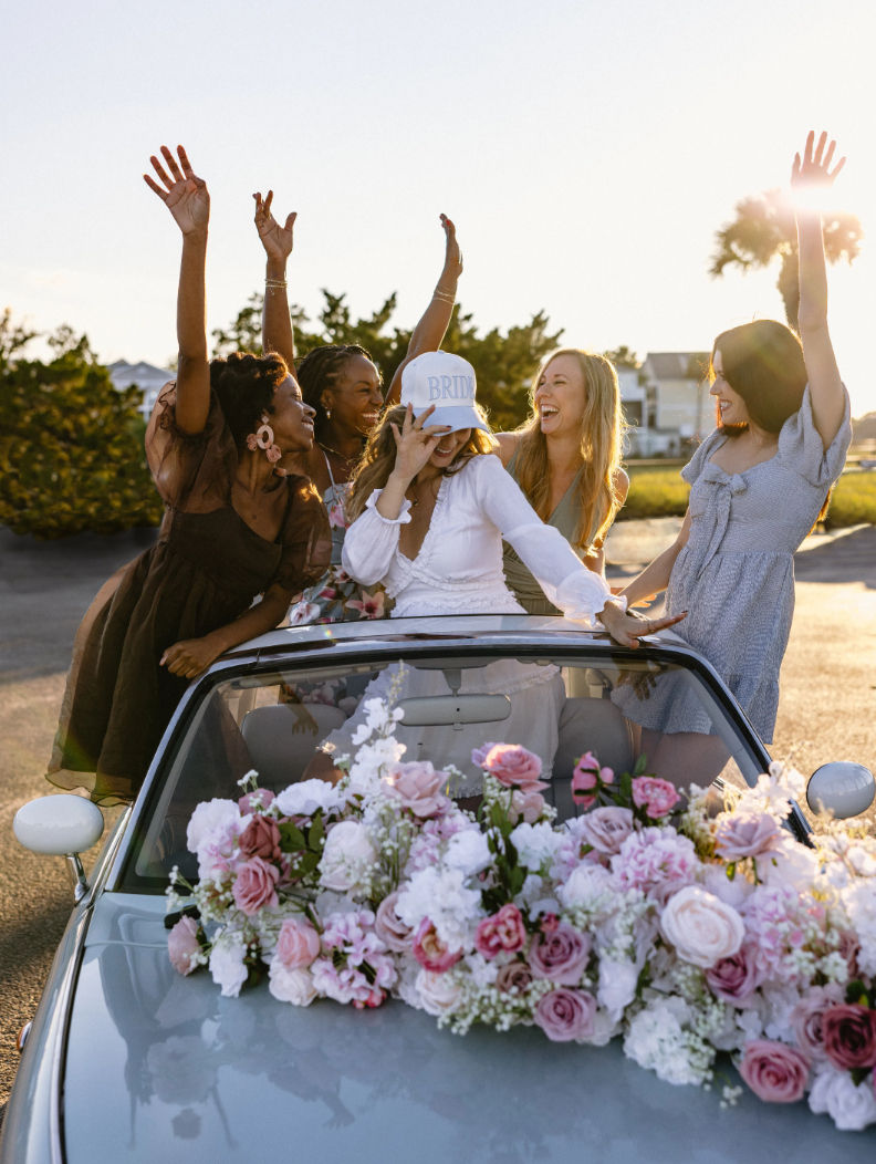Bachelorette party: bride-to-be in a 'BRIDE' cap and friends cheering in a flower-adorned convertible at sunset on a coastal road