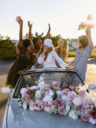 Bachelorette party: bride-to-be in a 'BRIDE' cap and friends cheering in a flower-adorned convertible at sunset on a coastal road