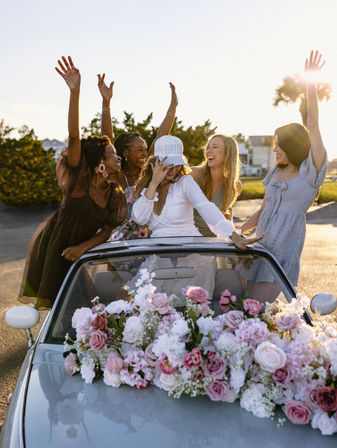 Bachelorette party: bride-to-be in a 'BRIDE' cap and friends cheering in a flower-adorned convertible at sunset on a coastal road