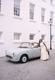 Stylish woman in a cream dress leaning on a pale-blue vintage convertible parked on a quiet street in front of a white colonial-style building with sash windows and a black lamppost.