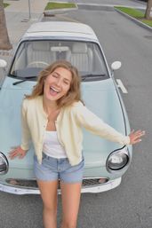 Laughing young woman in a cream cardigan and denim shorts leaning on a mint-green vintage car parked on a city street, sunny casual summer vibe