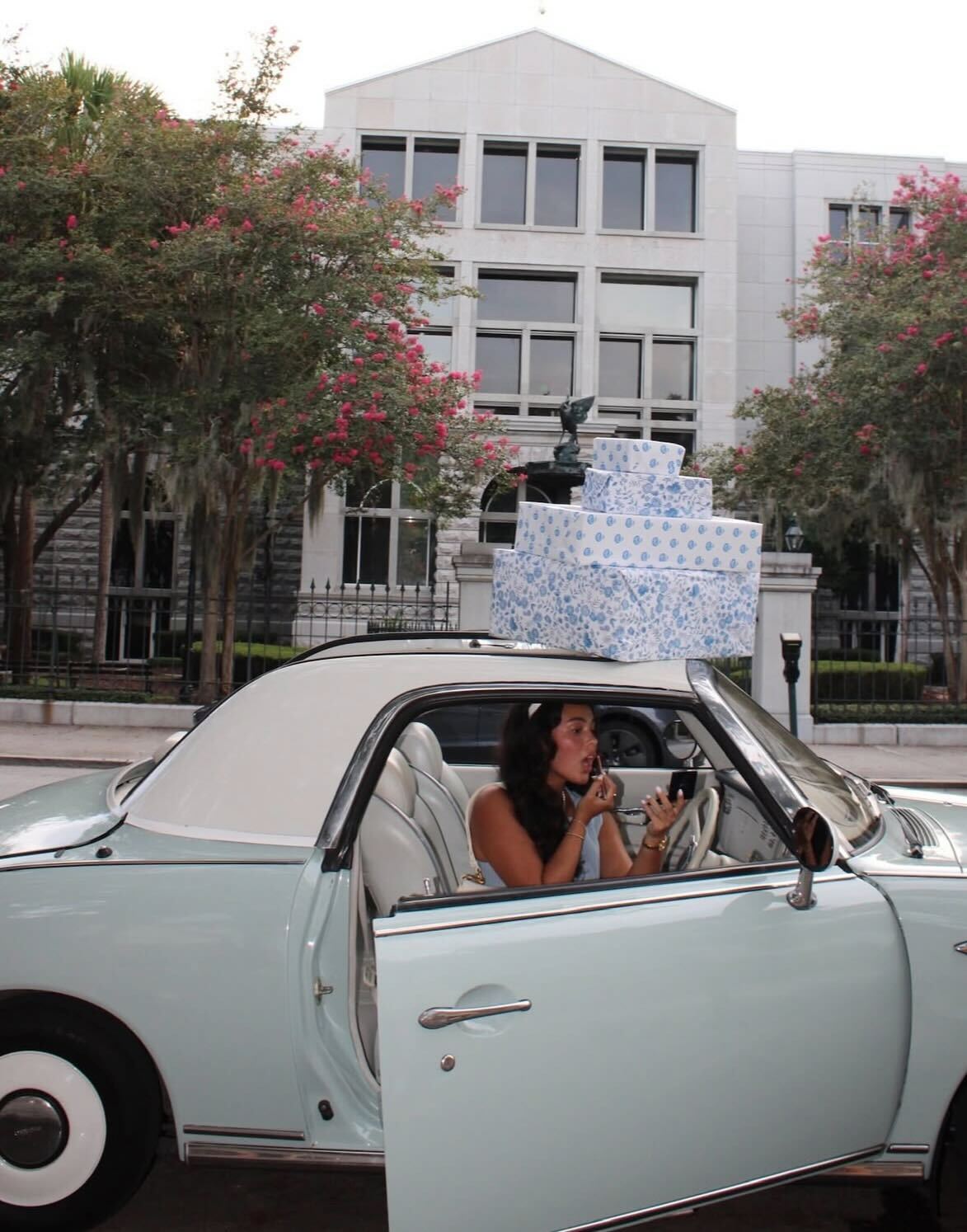 Woman applying lipstick inside a pastel blue vintage car with open door, three stacked blue-patterned gift boxes on the roof, parked on a tree-lined downtown street in front of a stone office building.