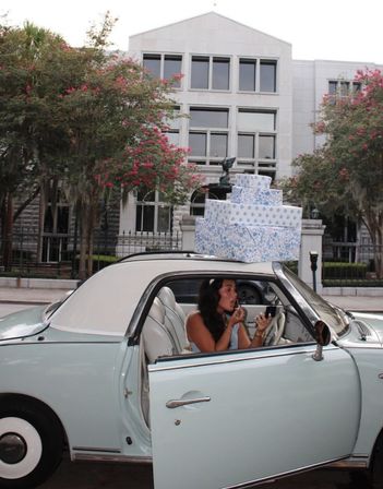 Woman applying lipstick inside a pastel blue vintage car with open door, three stacked blue-patterned gift boxes on the roof, parked on a tree-lined downtown street in front of a stone office building.